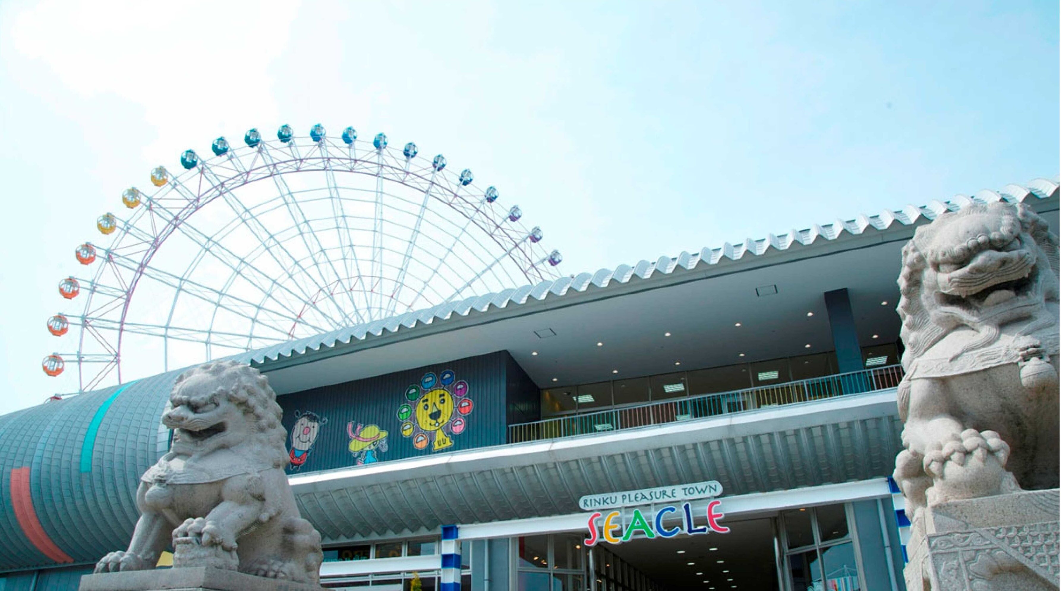 Un bâtiment avec une grande roue en arrière-plan, une enseigne indiquant « Rinku Pleasure Town », et deux statues de lions en pierre devant.