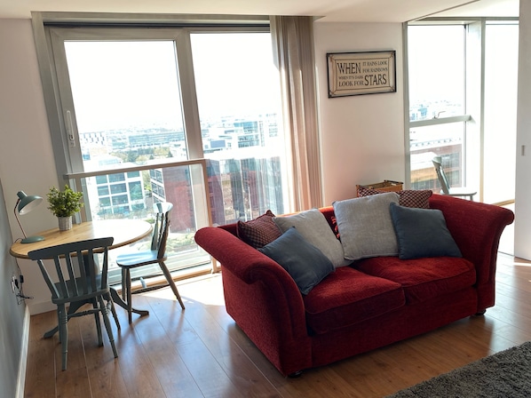 A living room with a red sofa, a small table with a lamp, and a view of the city through large windows.