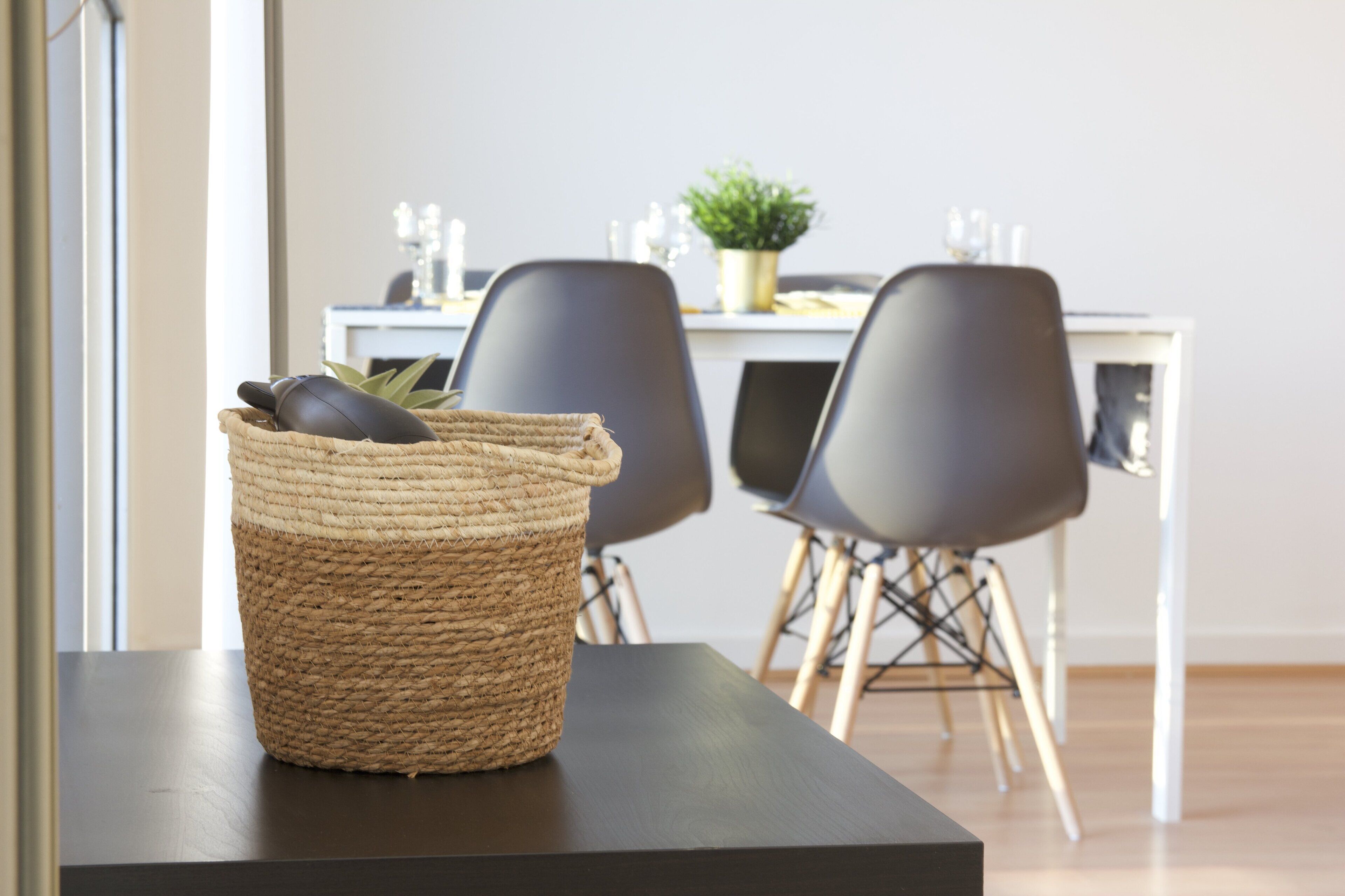 Una cesta tejida sobre una mesa de madera oscura con una planta en su interior, dos sillas grises y una mesa blanca con otra planta en maceta al fondo.