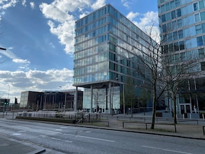 A modern building with a glass facade, a pedestrian walkway, and a street with a traffic light.