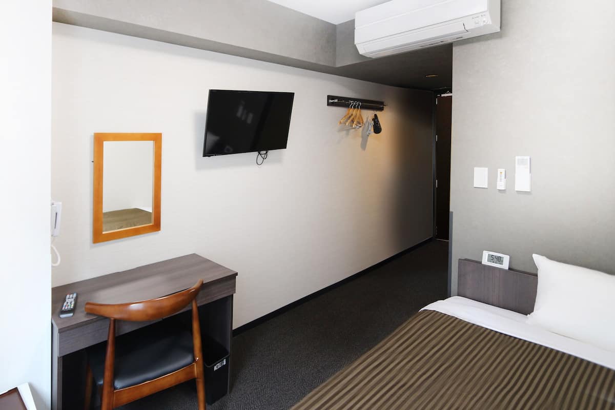Een hotelkamer met een bed, een bureau met stoel, een televisie, een spiegel en een kapstok met hangende spullen.
