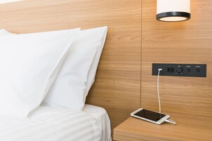 Una cama bien hecha con sábanas y almohadas blancas, una mesita de noche con un teléfono inteligente cargando y un enchufe de pared con puertos etiquetados.