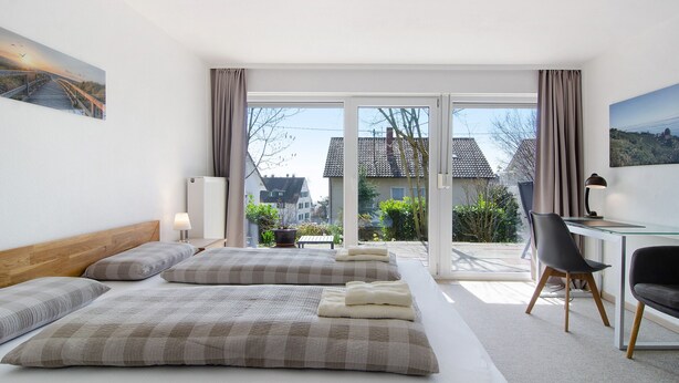 A hotel room with a bed, a desk, a chair, and a view of the outside. - Apartment 'Fewo Gartenterrasse' with Private Terrace, Garden, and Wi-Fi (Meersburg)