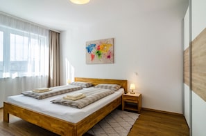 A neatly made bed with a wooden headboard, a bedside table with a lamp, a colorful abstract painting on the wall, and a large window with sheer curtains.