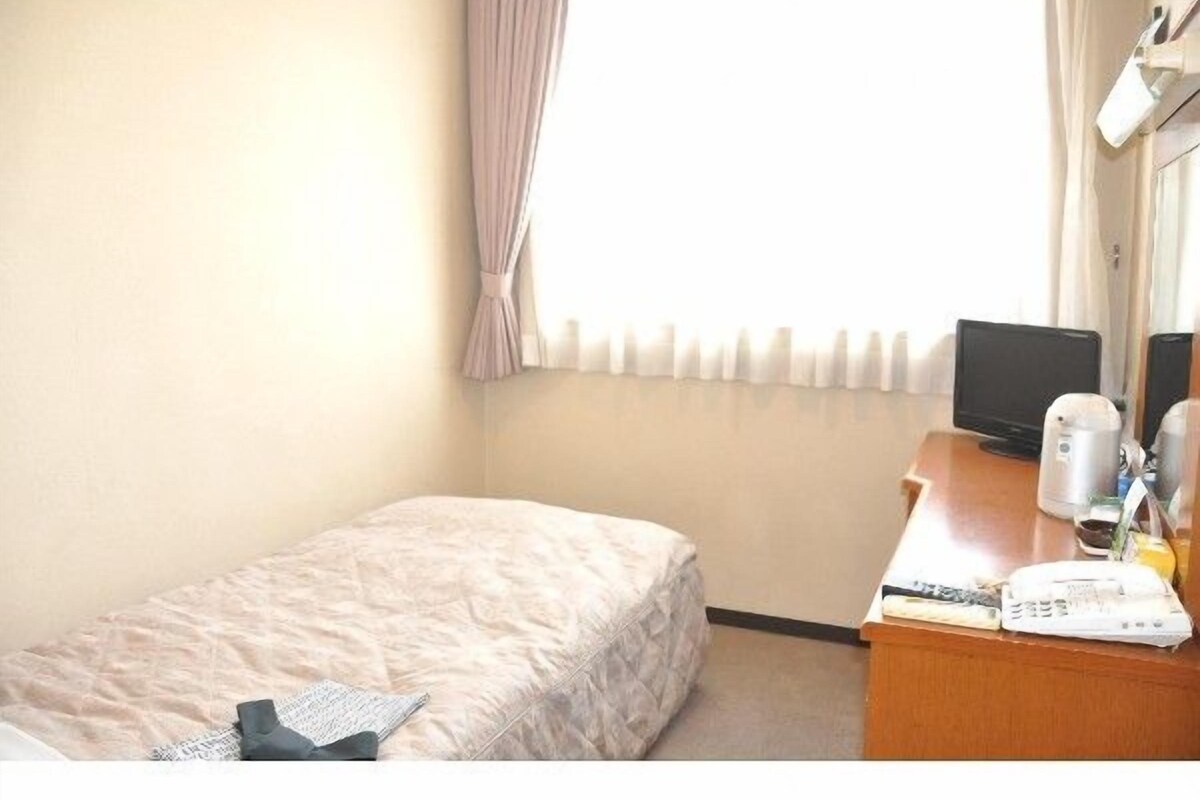 A hotel room with a single bed, a desk with a computer, a kettle, and a telephone.
