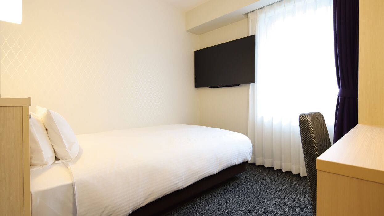 Habitación de hotel con cama, escritorio, silla, televisor montado en la pared y ventana con cortinas.
