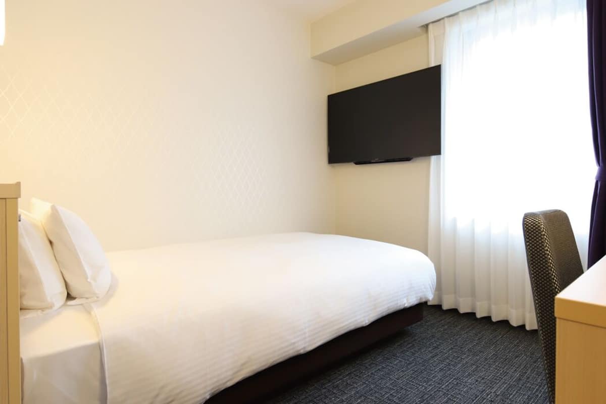 Ein Hotelzimmer mit einem Bett, einem Schreibtisch, einem Sessel, einem an der Wand befestigten Fernseher und einem Fenster mit Vorhängen.