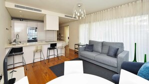 Ein modernes Wohnzimmer mit grauem Sofa, einem Esstisch mit Stühlen, einer Küchenzeile mit weißer Arbeitsplatte und Edelstahlgeräten sowie einem großen Fenster mit durchsichtigen Vorhängen.