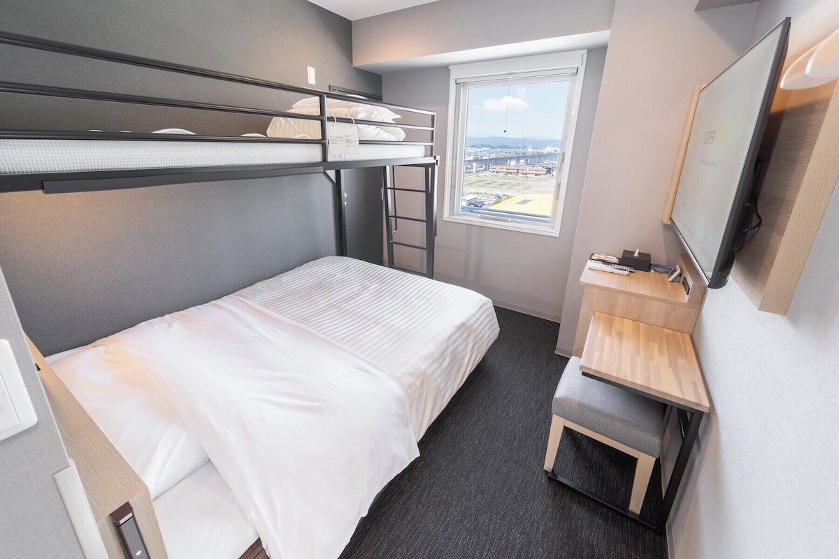 Ein kleines Hotelzimmer mit Etagenbetten, einem Schreibtisch, einem Stuhl, einem Fernseher und einem Fenster mit Blick auf die Stadt.