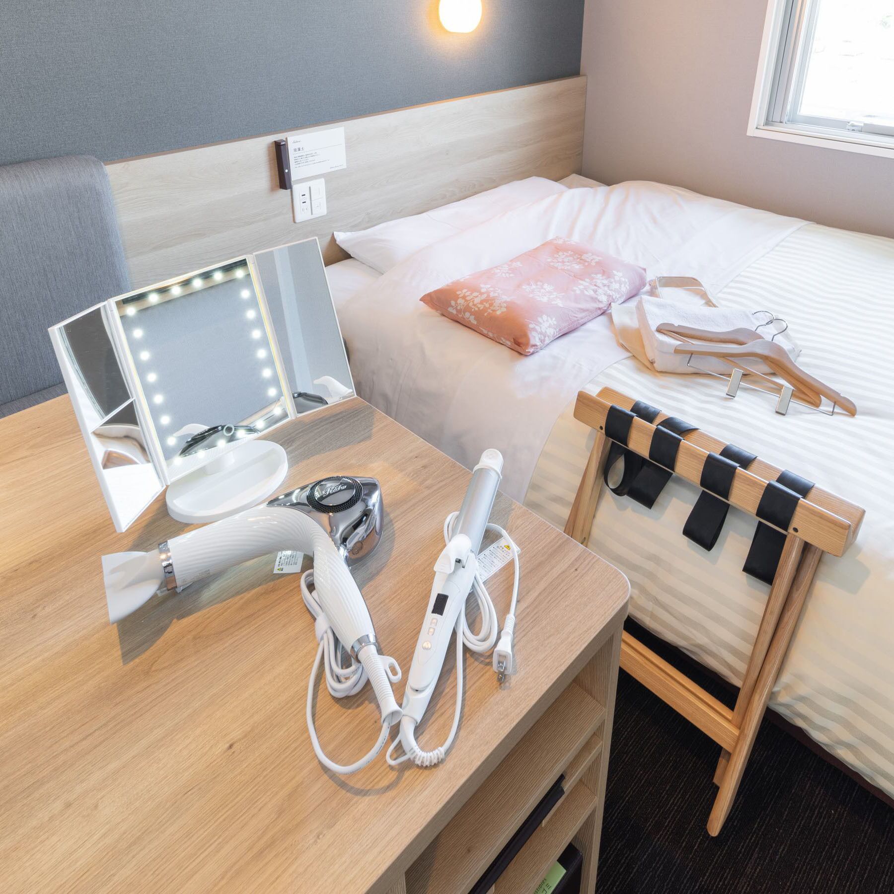 Une chambre d’hôtel comprenant un lit, une coiffeuse avec un miroir et une table de chevet en bois, ainsi qu’un sèche-cheveux et d’autres articles de toilette.