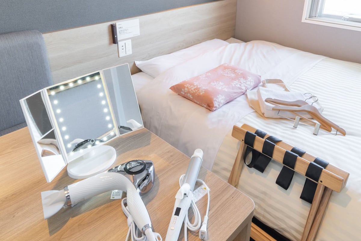 Ein Hotelzimmer mit einem Bett, einem Schminktisch mit Spiegel und einem Holztischchen mit Fön und weiteren Pflegeprodukten.