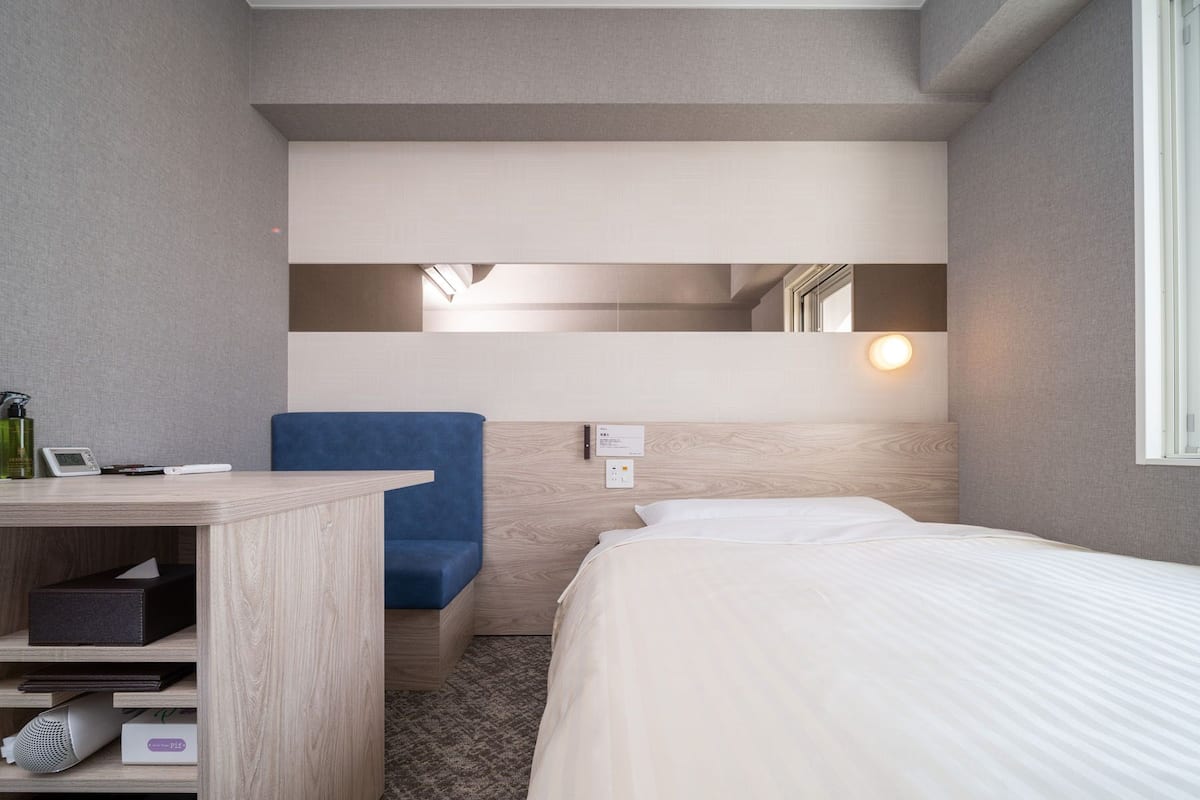 Ein kleines Hotelzimmer mit Bett, Schreibtisch, Stuhl, Spiegel und einem kleinen Regal mit Gegenständen.