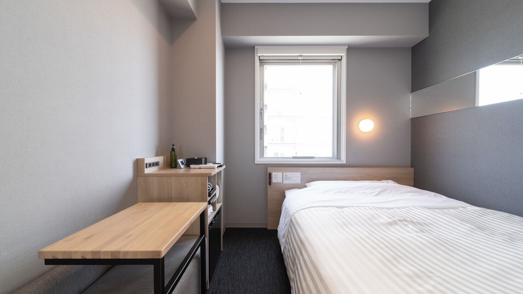 Une chambre d’hôtel de taille réduite, comprenant un lit simple, un bureau, une petite table, une fenêtre avec vue et un luminaire fixé au mur.