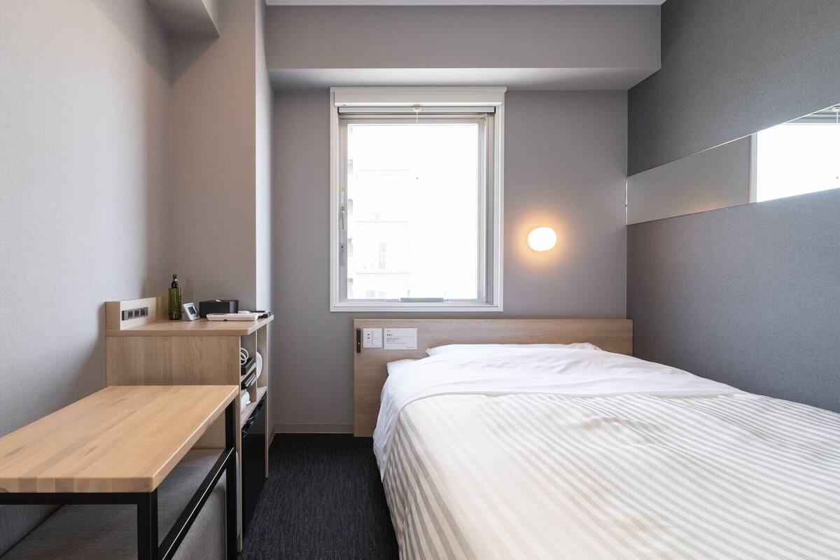 Une chambre d’hôtel de taille réduite, comprenant un lit simple, un bureau, une petite table, une fenêtre avec vue et un luminaire fixé au mur.