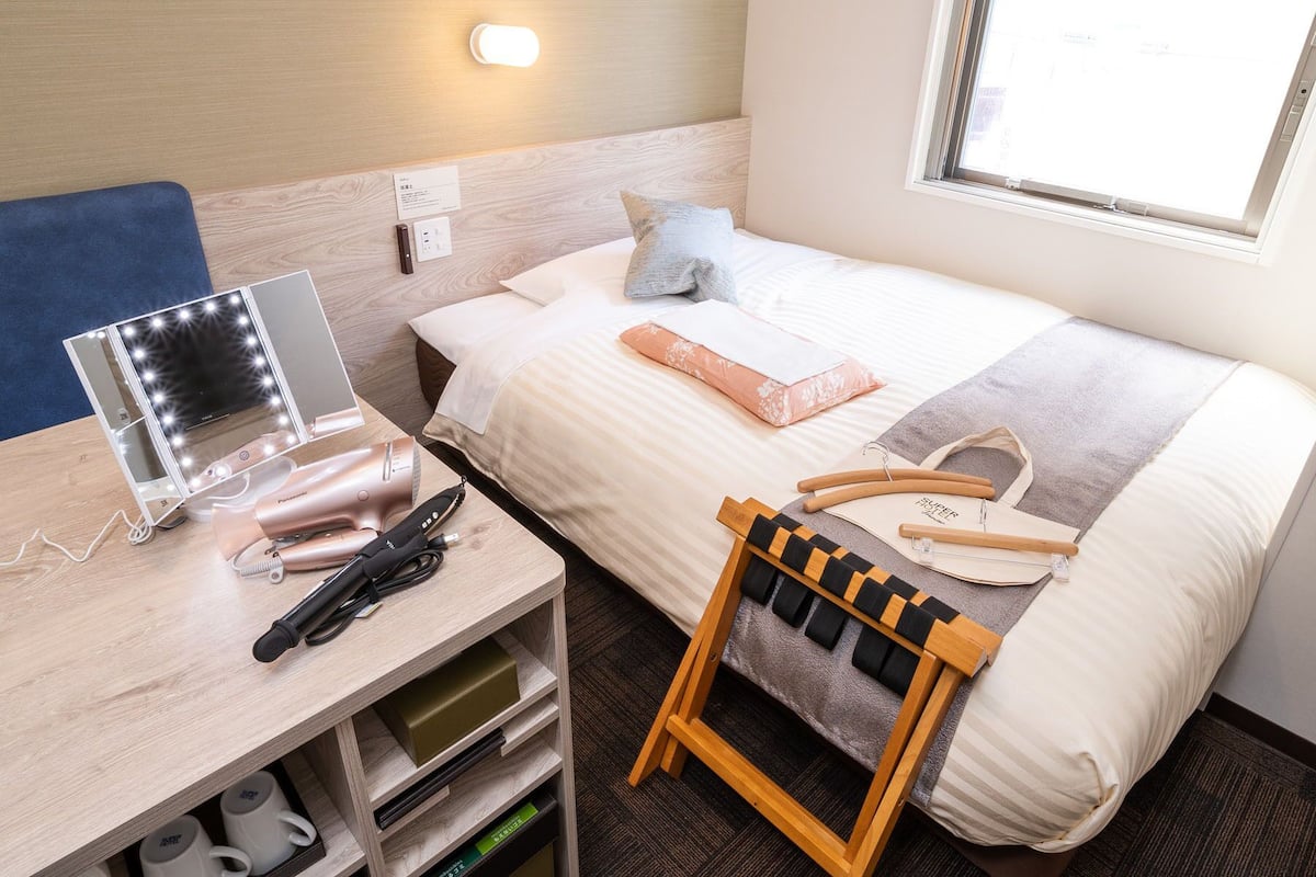 Ein Hotelzimmer mit Bett, Schreibtisch mit Schminkspiegel, Schuhregal und Fenster.