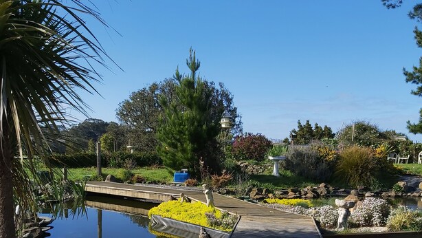A wooden deck leading to a pond with a statue, surrounded by greenery and a clear blue sky. - Kingfisher Cabin. Nestled Pukekohe side of Bombay, with a large private pool. (Bombay)