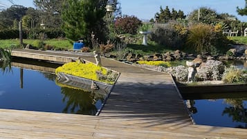 A wooden deck leading to a pond with a statue, surrounded by greenery and a clear blue sky.