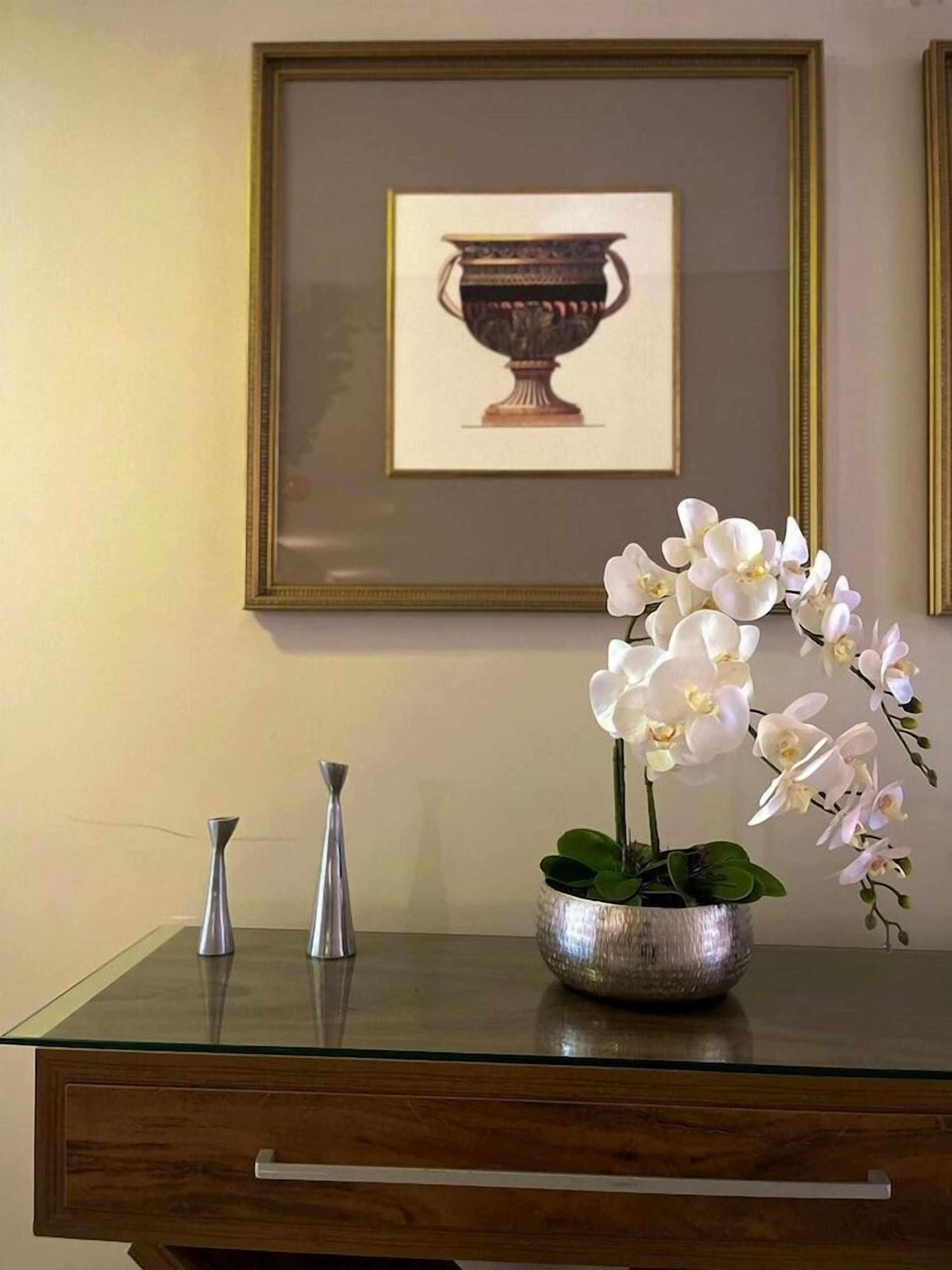 Een glazen tafel met een metalen vaas en een ingelijste foto aan de muur.