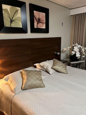 Una habitación de hotel con una cama grande, dos cuadros con motivos florales en la pared y un florero con flores blancas sobre una mesita.