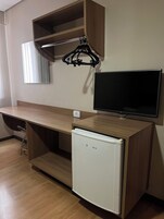Quarto compacto com escrivaninha de madeira, televisão e um pequeno frigorífico.