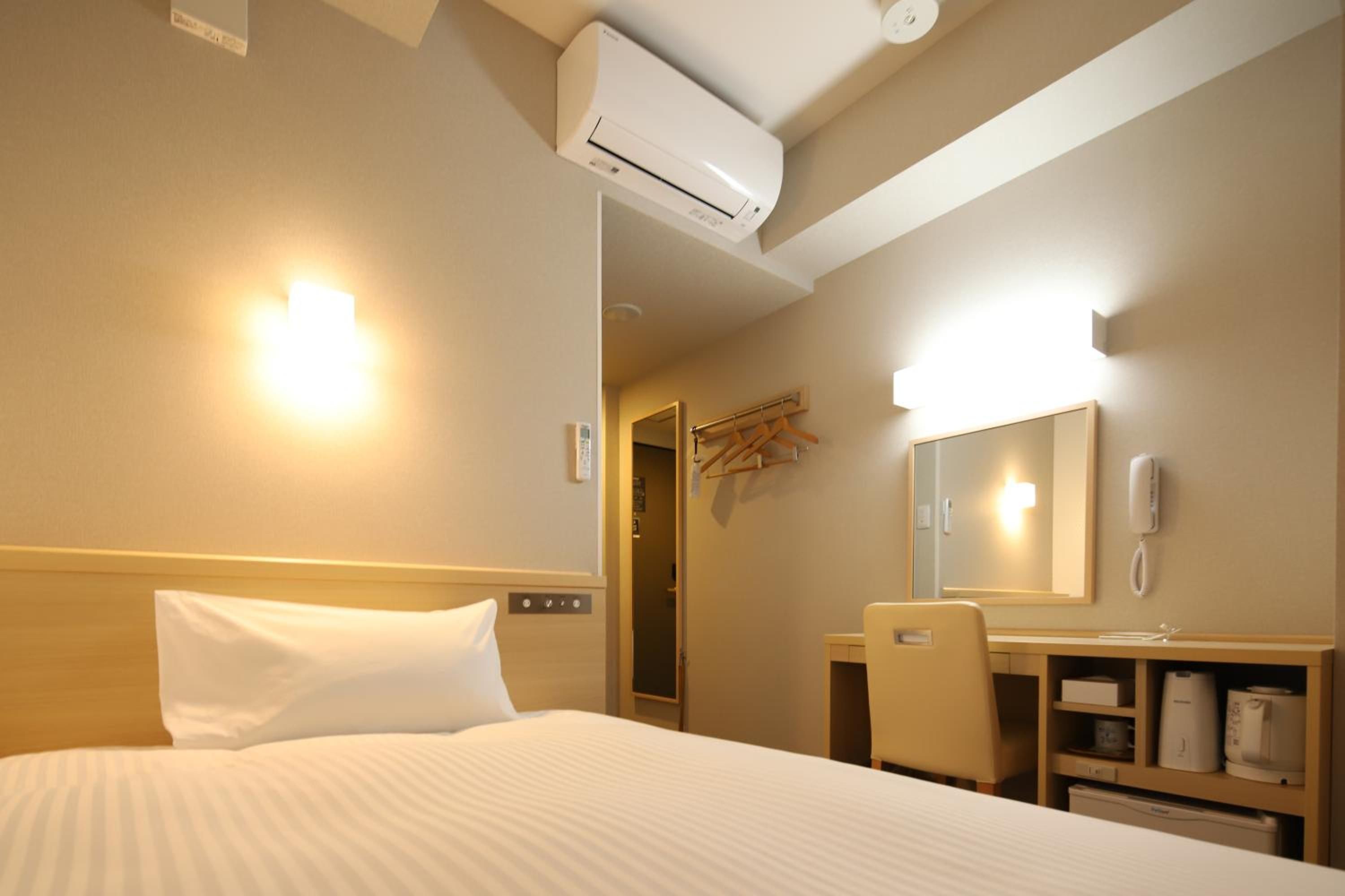 A hotel room with a bed, a desk, a chair, a mirror, and an air conditioning unit.