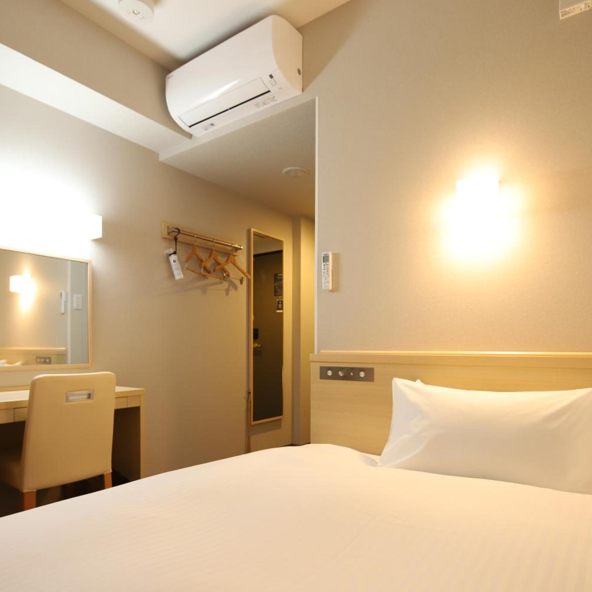 Una habitación de hotel con una cama, un escritorio, una silla, un espejo, un equipo de aire acondicionado, una lámpara de pared y una puerta.