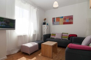 A modern living room with a grey sofa, a wooden coffee table, and a pink ottoman. A flat-screen TV is mounted on the wall, and there is a white cabinet with a vase on top.