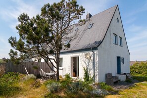 Une maison blanche avec un toit en pente, une terrasse en bois et un grand arbre à l’avant.