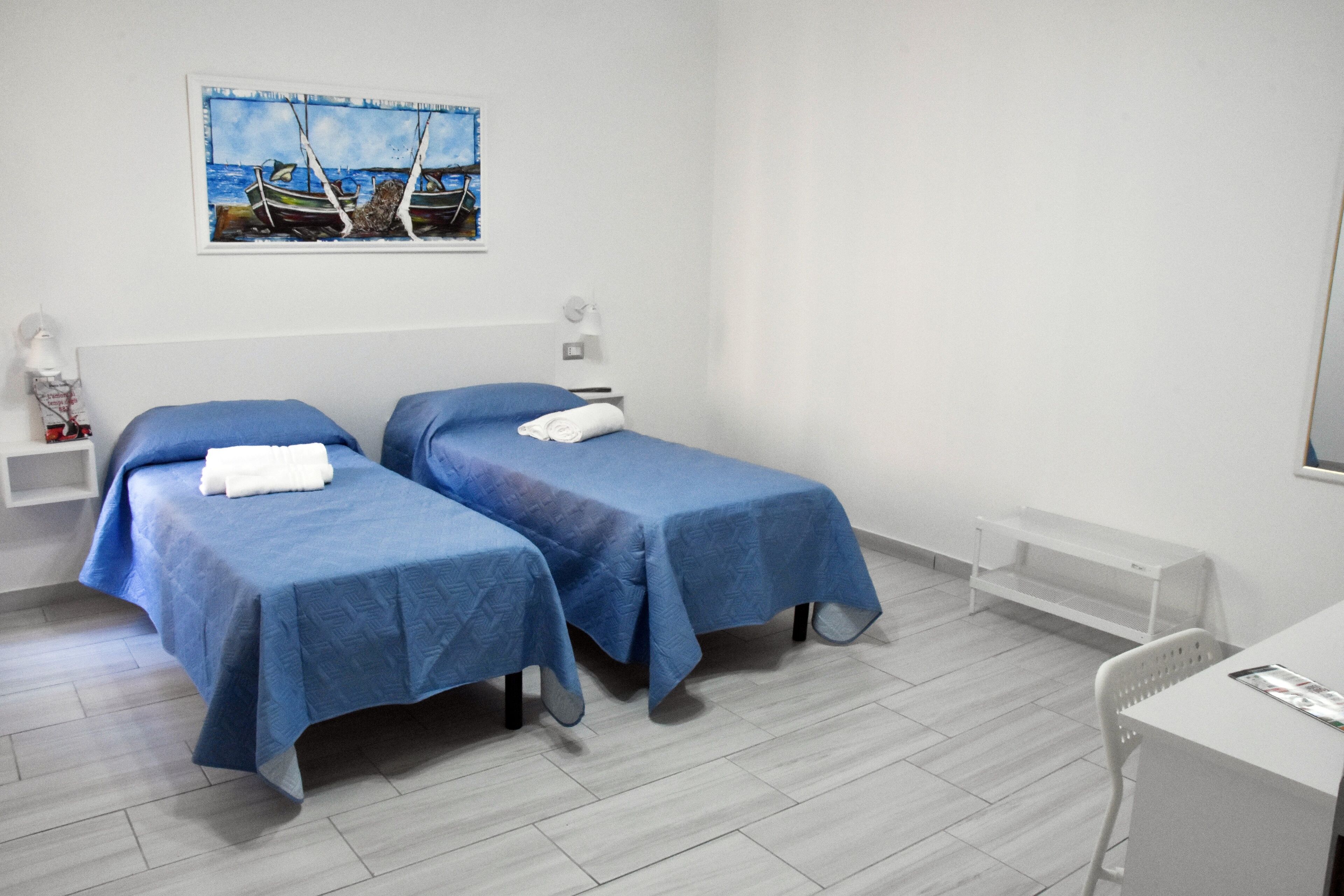A hotel room with two beds, a desk, a chair, a small shelf, a bedside table, a lamp, and a painting of boats on the wall.
