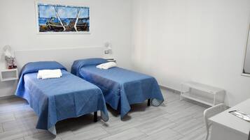A hotel room with two beds, a desk, a chair, a small shelf, a bedside table, a lamp, and a painting of boats on the wall.