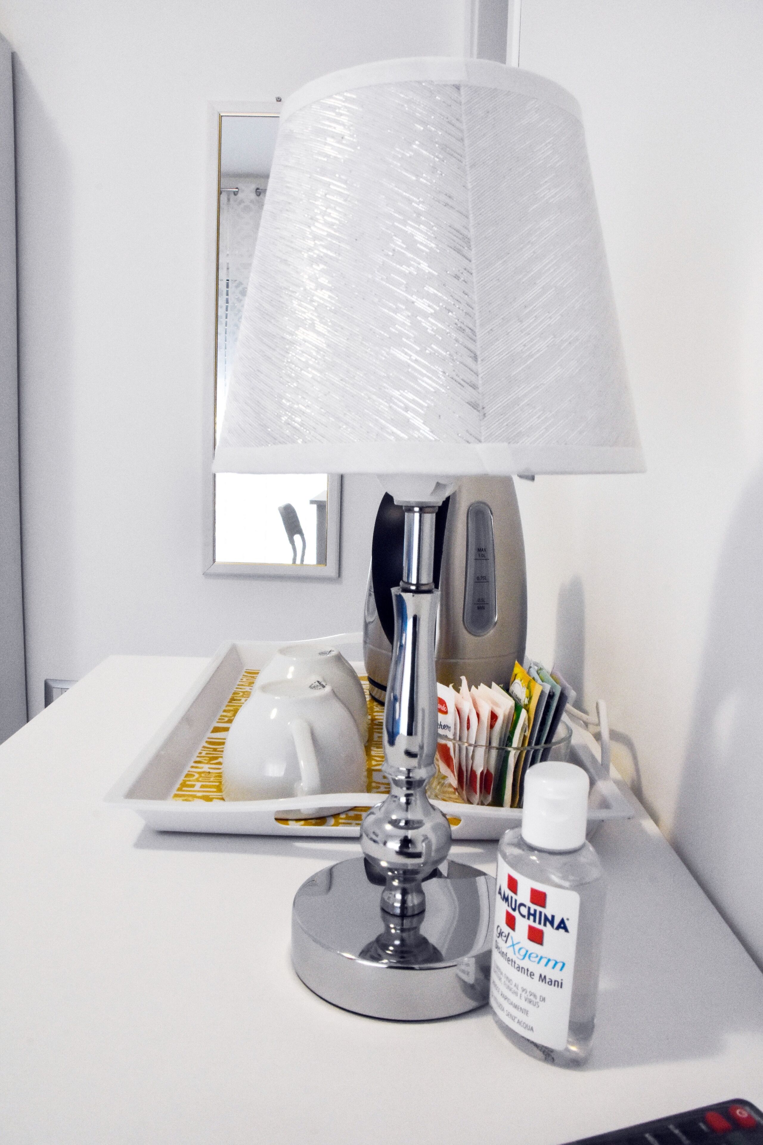 A hotel room with a white table, a lamp with a white lampshade, a teapot, a bottle of hand sanitizer, and a tray with various items.