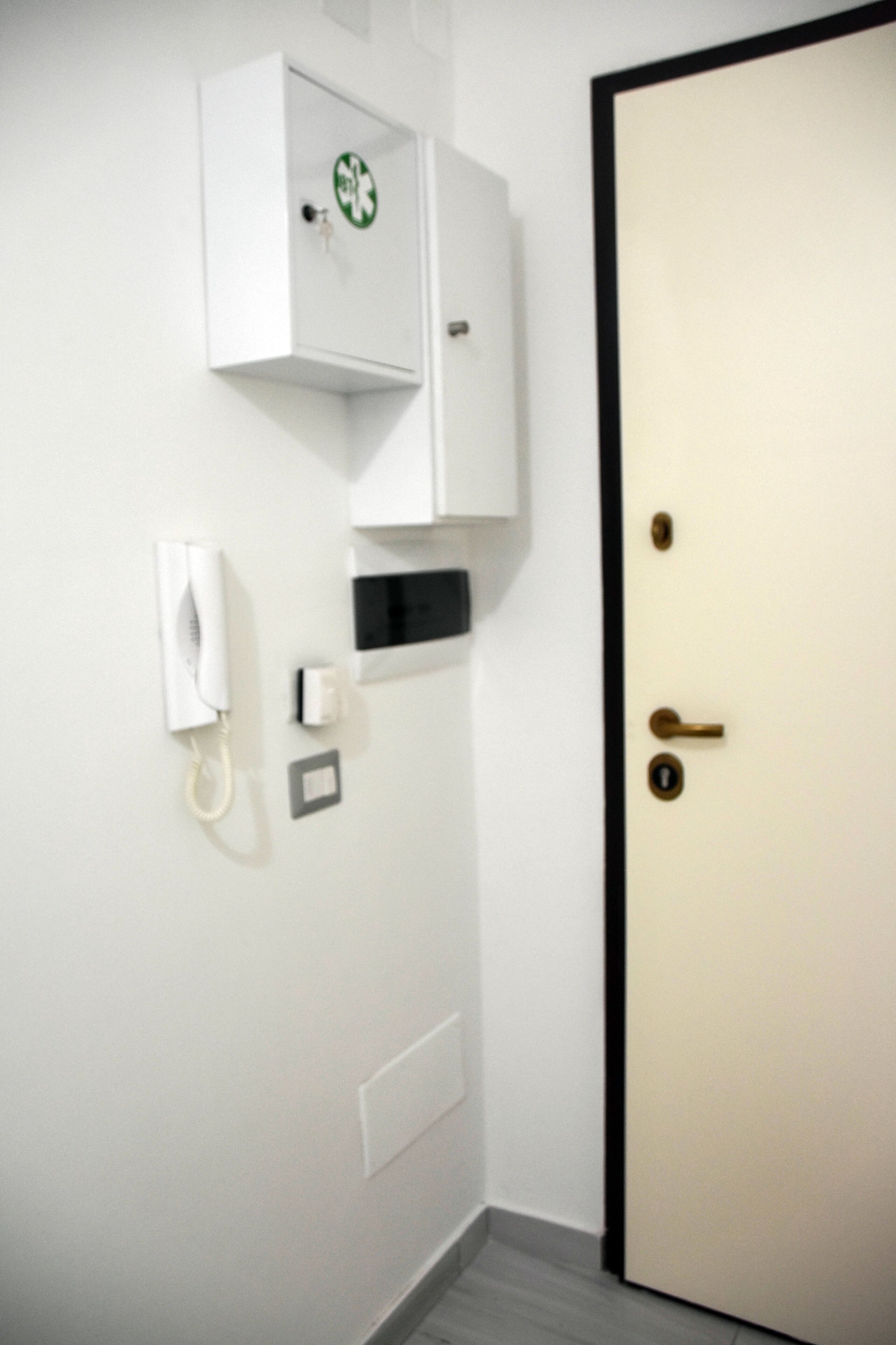 Una habitación con una puerta blanca, un armario blanco con un logotipo verde, un teléfono blanco y un interruptor de luz en la pared.