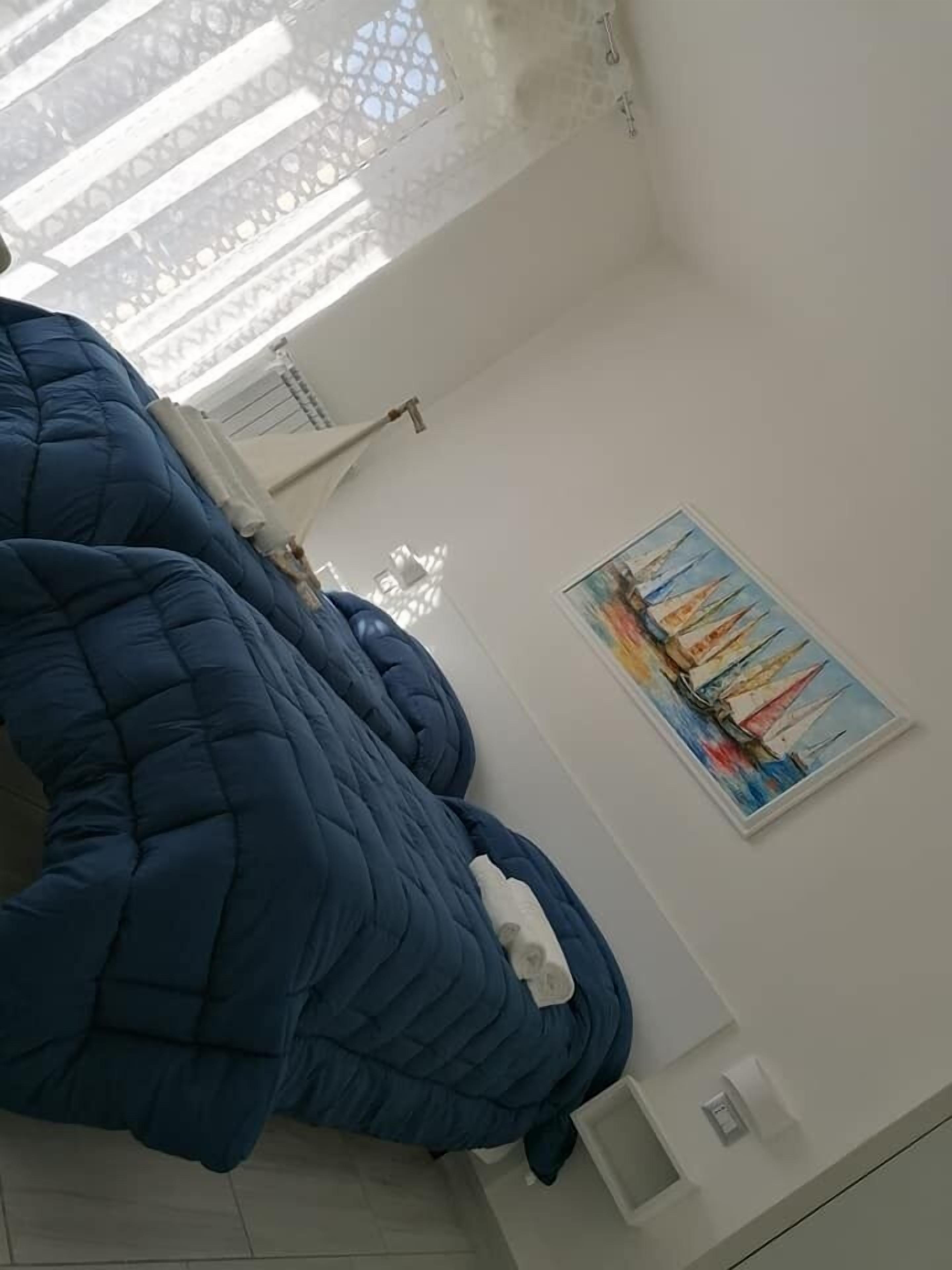 Ein Zimmer mit einem großen, blauen Bett, einem Gemälde an der Wand und einem Fenster mit weißem Vorhang.
