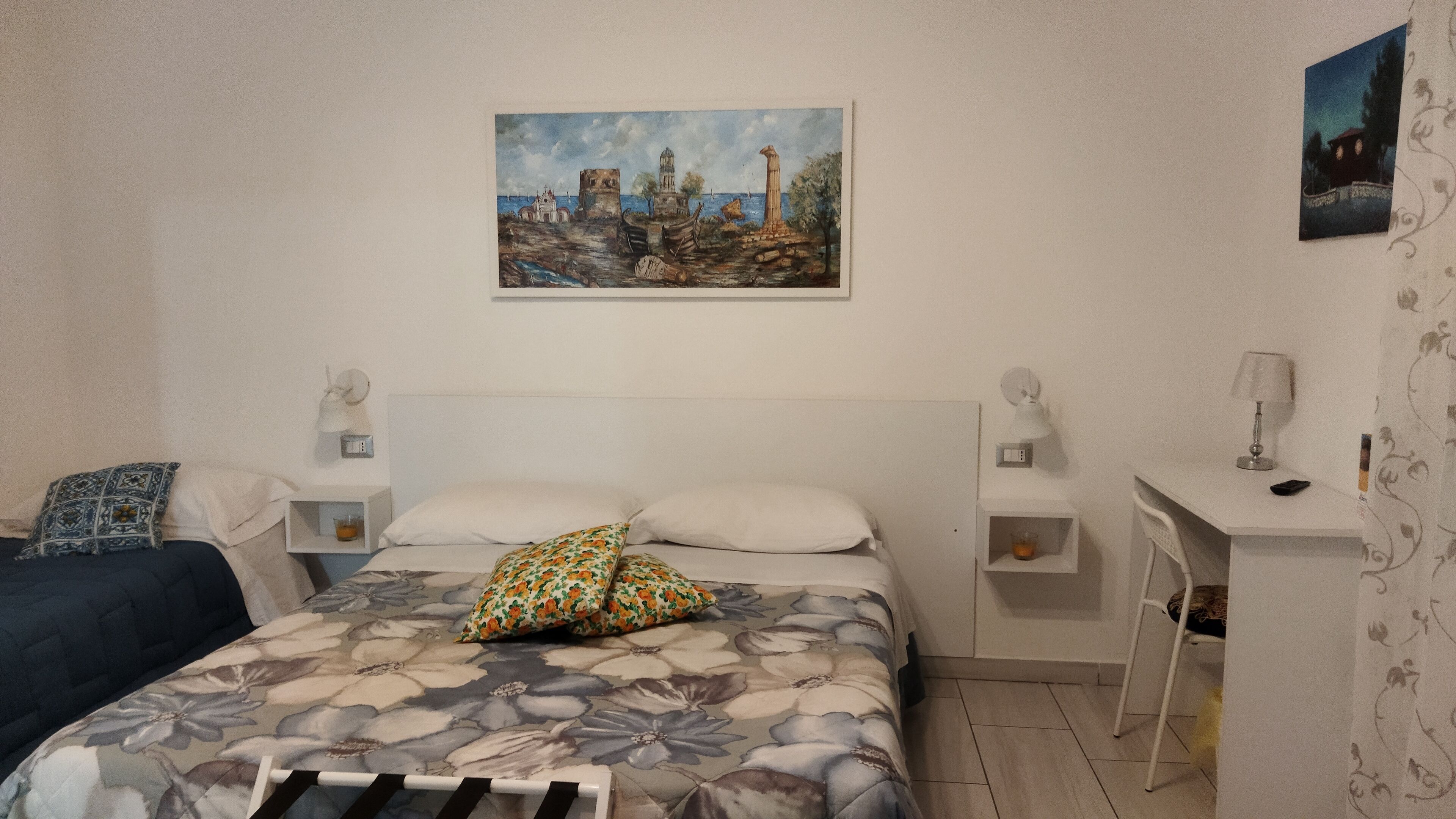 A bedroom with a bed, a nightstand, a desk, and a painting on the wall.