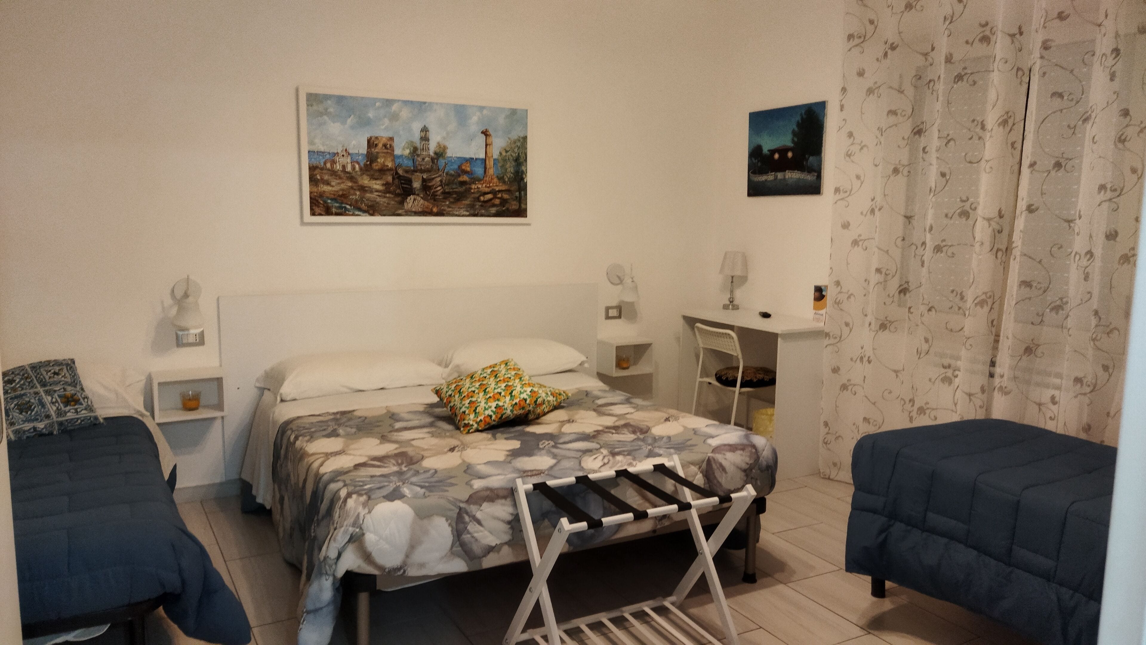 A bedroom with a bed, a desk, and two chairs. There are two paintings on the wall.