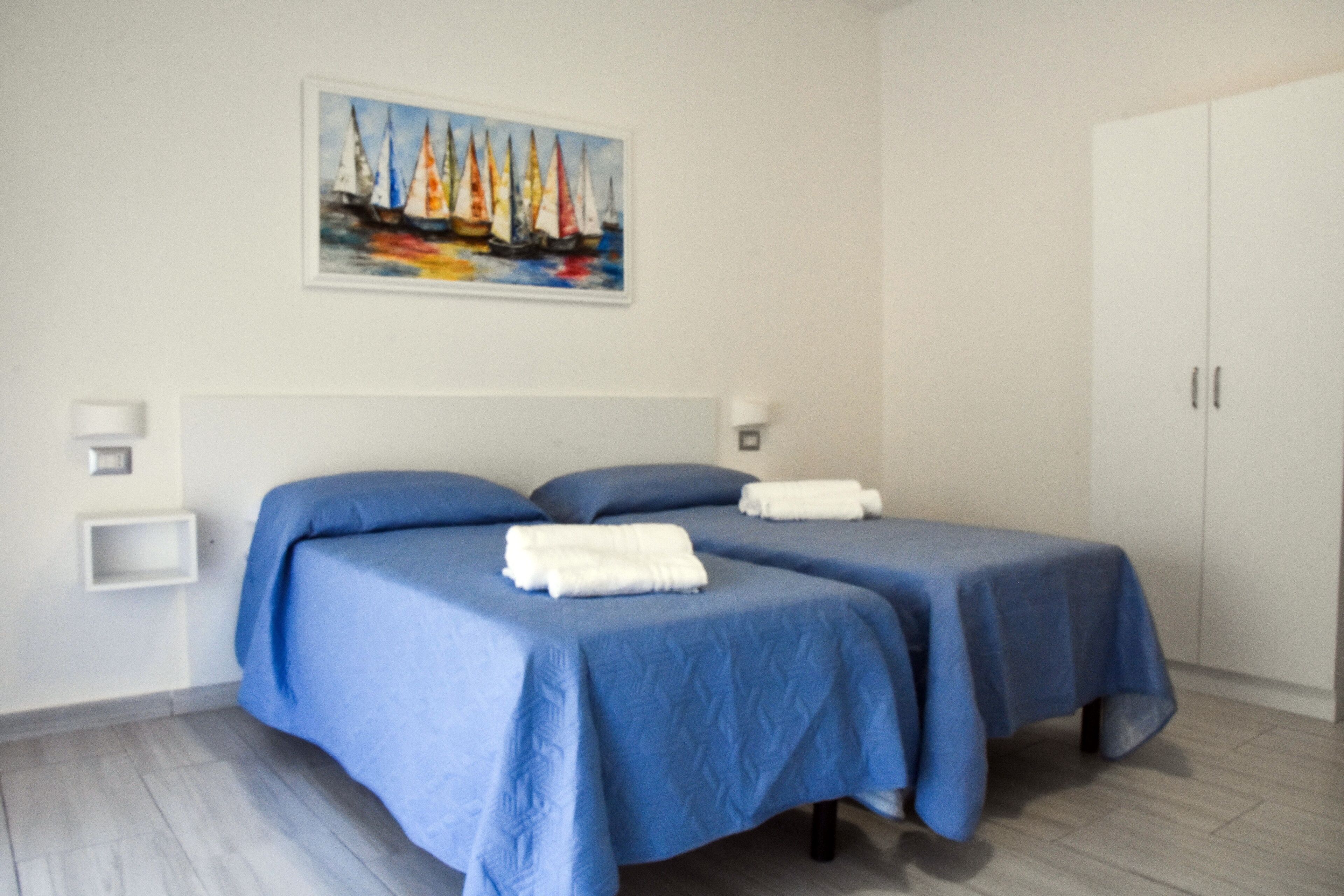 A room with two beds, a painting of sailboats on the wall, a white wardrobe, and a small shelf with a light.