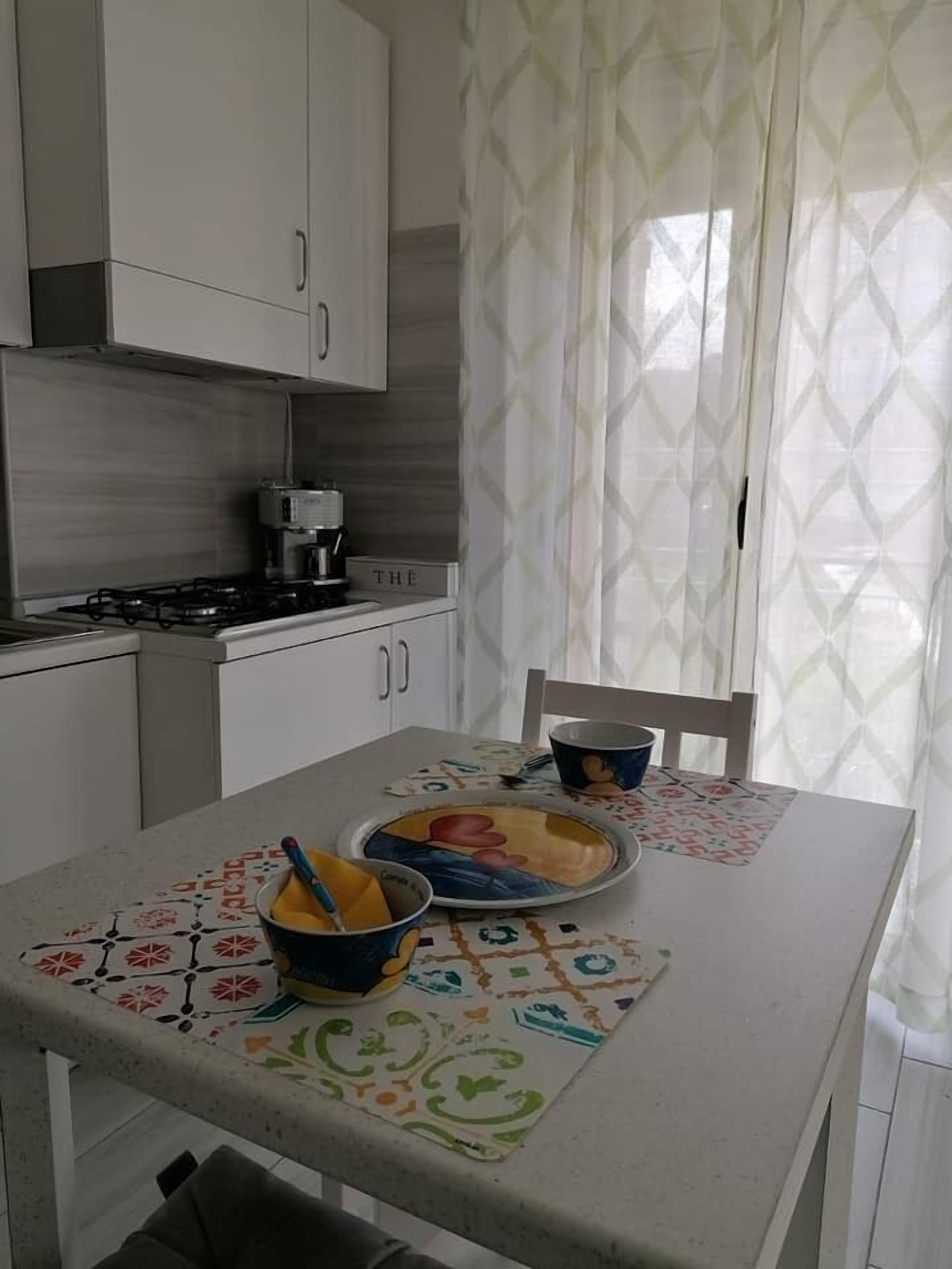 Una cocina con armarios blancos, una estufa y una cafetera. Hay una mesa con un mantel individual de colores y un bol con utensilios.