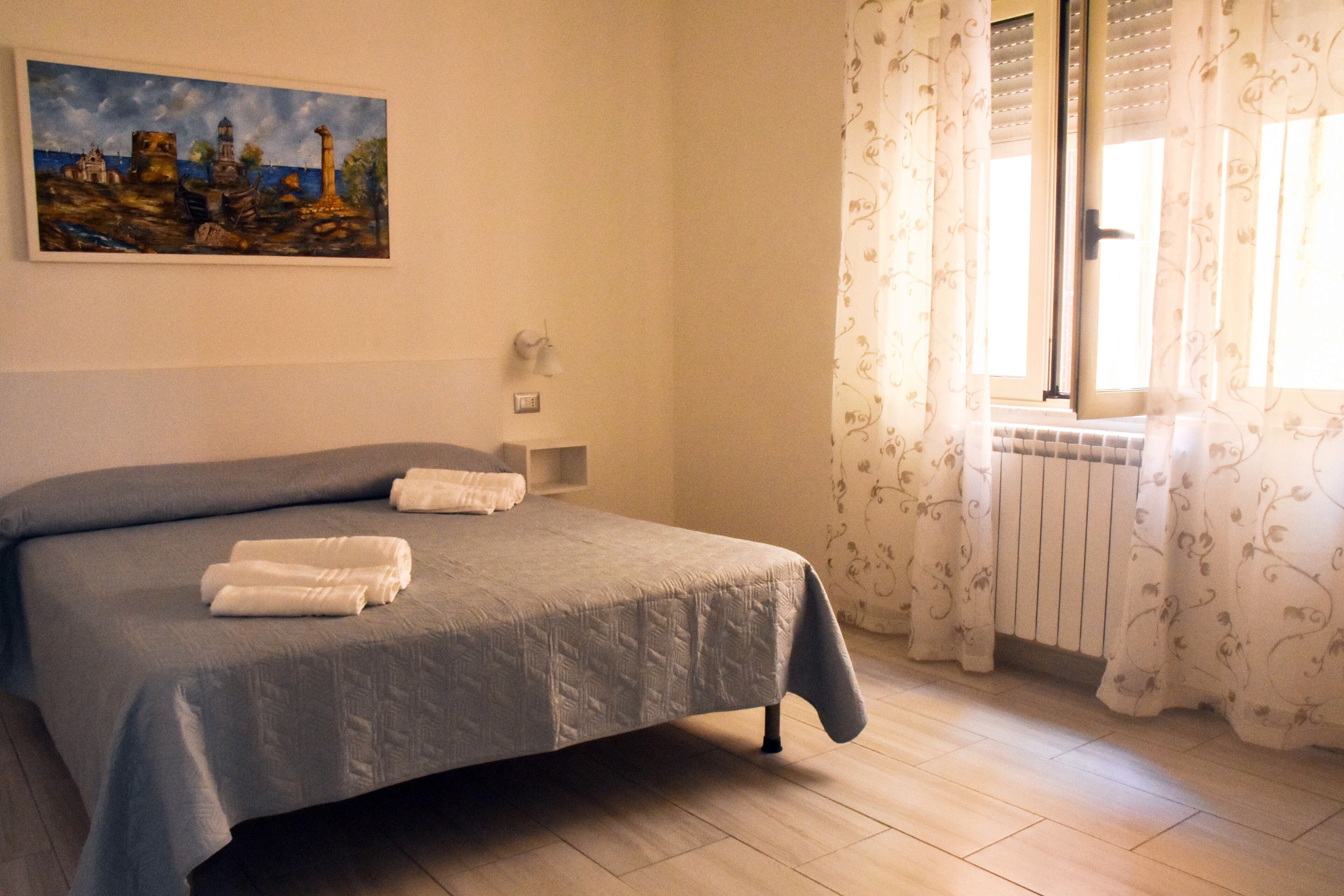 Een slaapkamer met een bed, een schilderij aan de muur, een raam met gordijnen en een radiator.
