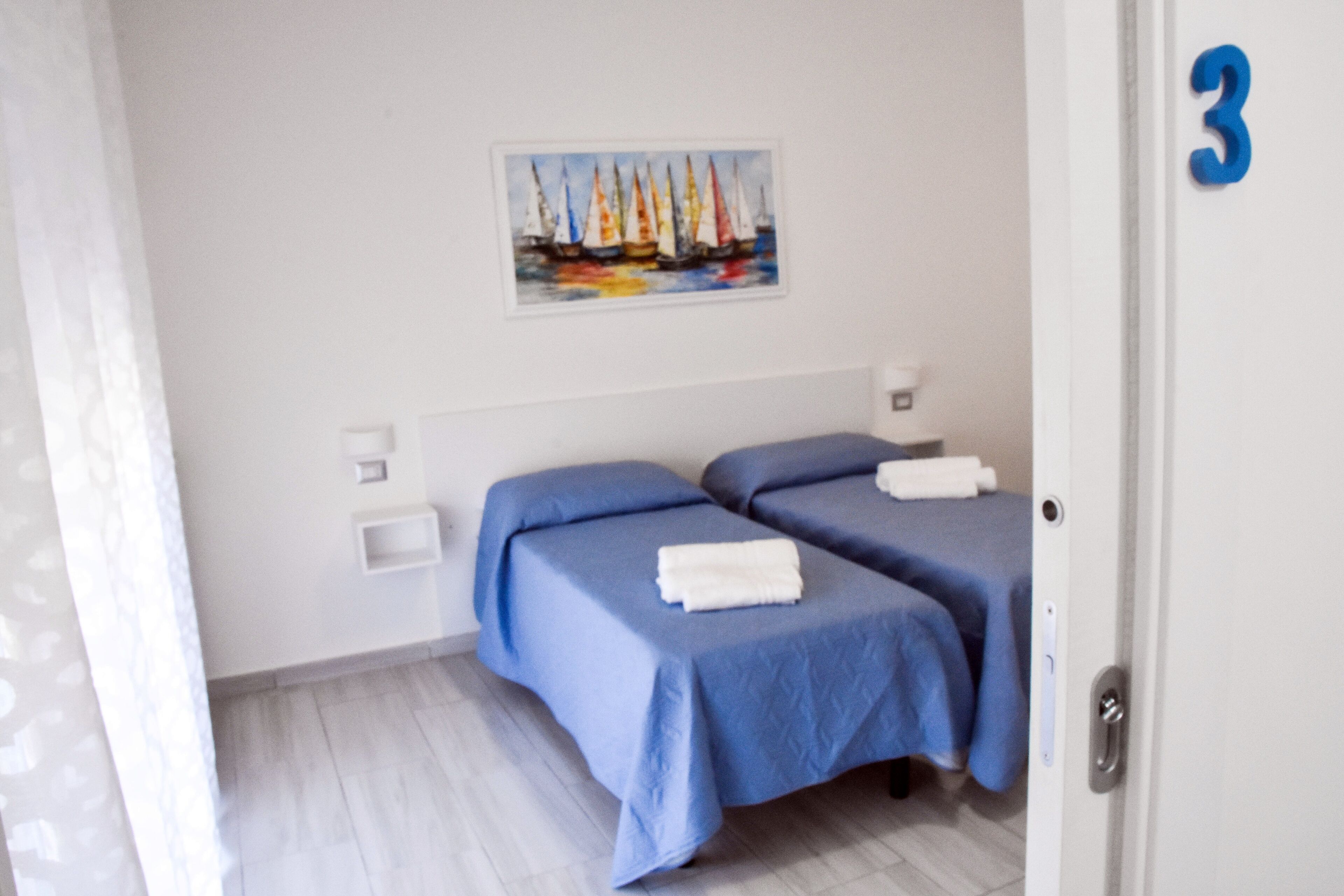 Een hotelkamer met twee bedden, een schilderij van zeilboten aan de muur en een deur met het nummer 3.