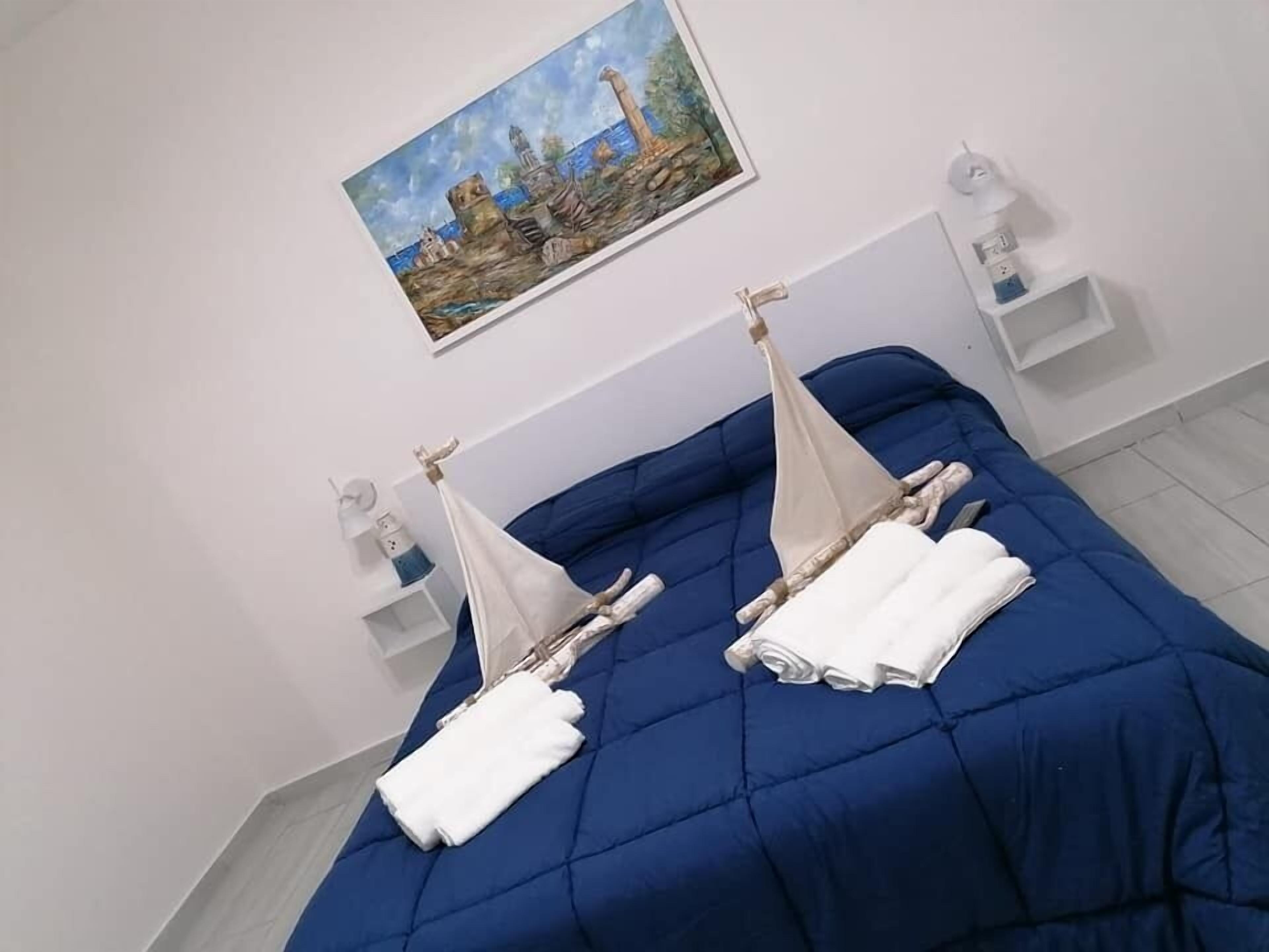 A room with a blue bedspread, two white towels, a painting of a cityscape, and a small white shelf with a lamp.