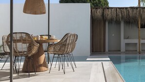 Un área junto a la piscina con sillas de mimbre, una mesa de madera y un techo de paja colgante.