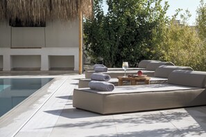 A poolside area with a modern lounge, a thatched roof, and a view of greenery.
