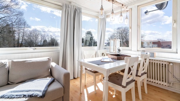Um quarto acolhedor com sofá, uma mesa de jantar com cadeiras e amplas janelas com vista para árvores e edifícios.