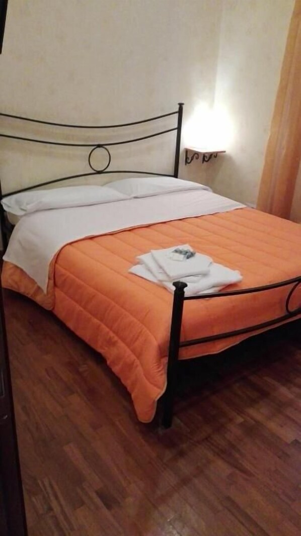 A single bed with an orange bedspread and white sheets, a black metal frame, and a small bedside table with a lamp.