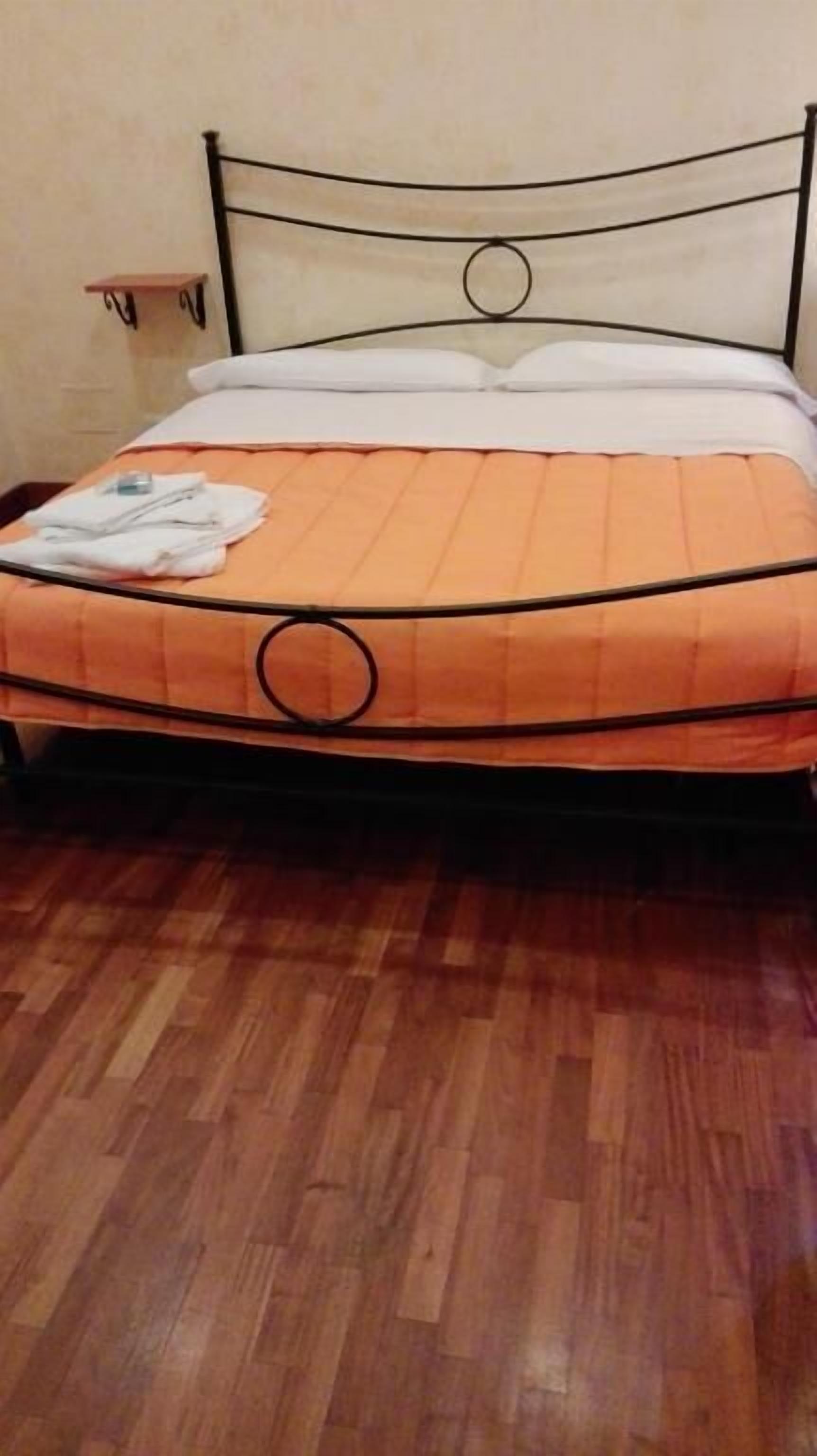 A bed with an orange mattress and a metal frame, a wooden floor, and a small bedside table with a lamp.
