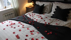 Una cama con pétalos de rosa esparcidos sobre las sábanas, un plaid de punto oscuro y una ventana con un pájaro posado en el alféizar.