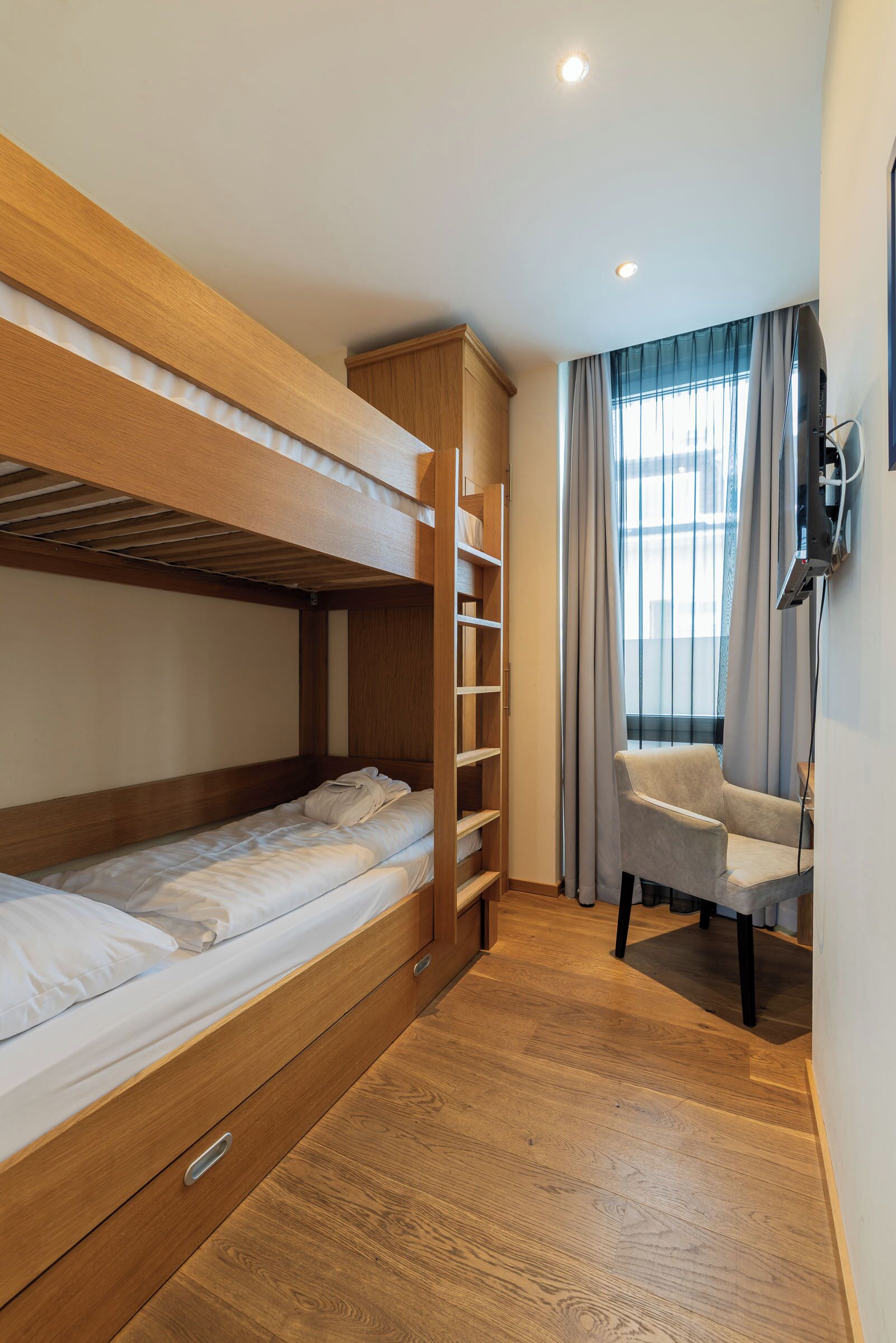 Une chambre avec un lit superposé, un plancher en bois, une chaise et une fenêtre avec des rideaux.