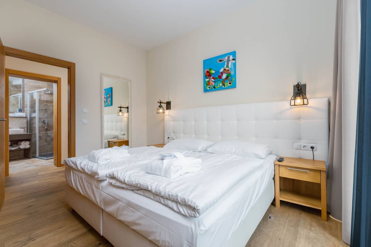 A modern hotel room with a large bed, a wooden nightstand, a wall-mounted light, and a painting above the bed.