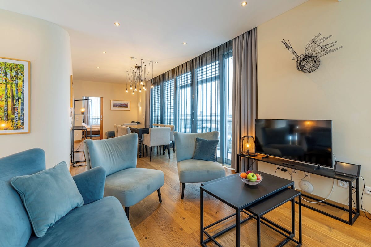 A modern living room with a sofa, armchairs, a coffee table, a TV, and a dining area in the background.