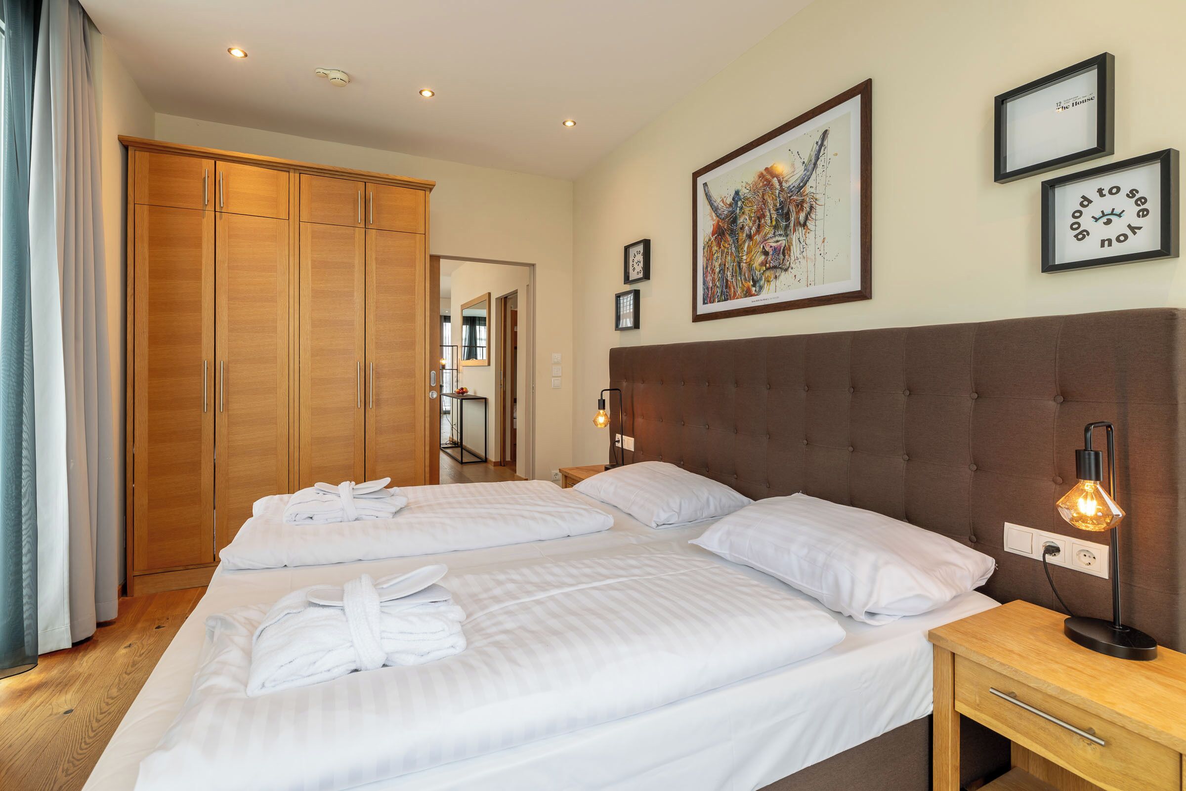 A hotel room with two beds, a large wardrobe, a headboard with a light fixture, and framed artwork on the wall.